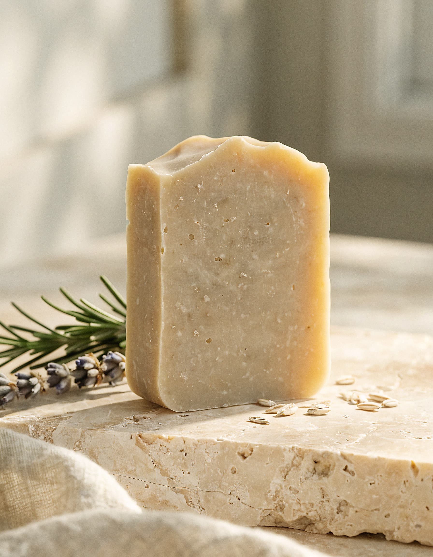 A handmade cold-process soap bar resting on travertine stone with rosemary, dried lavender, and oats in soft morning light.