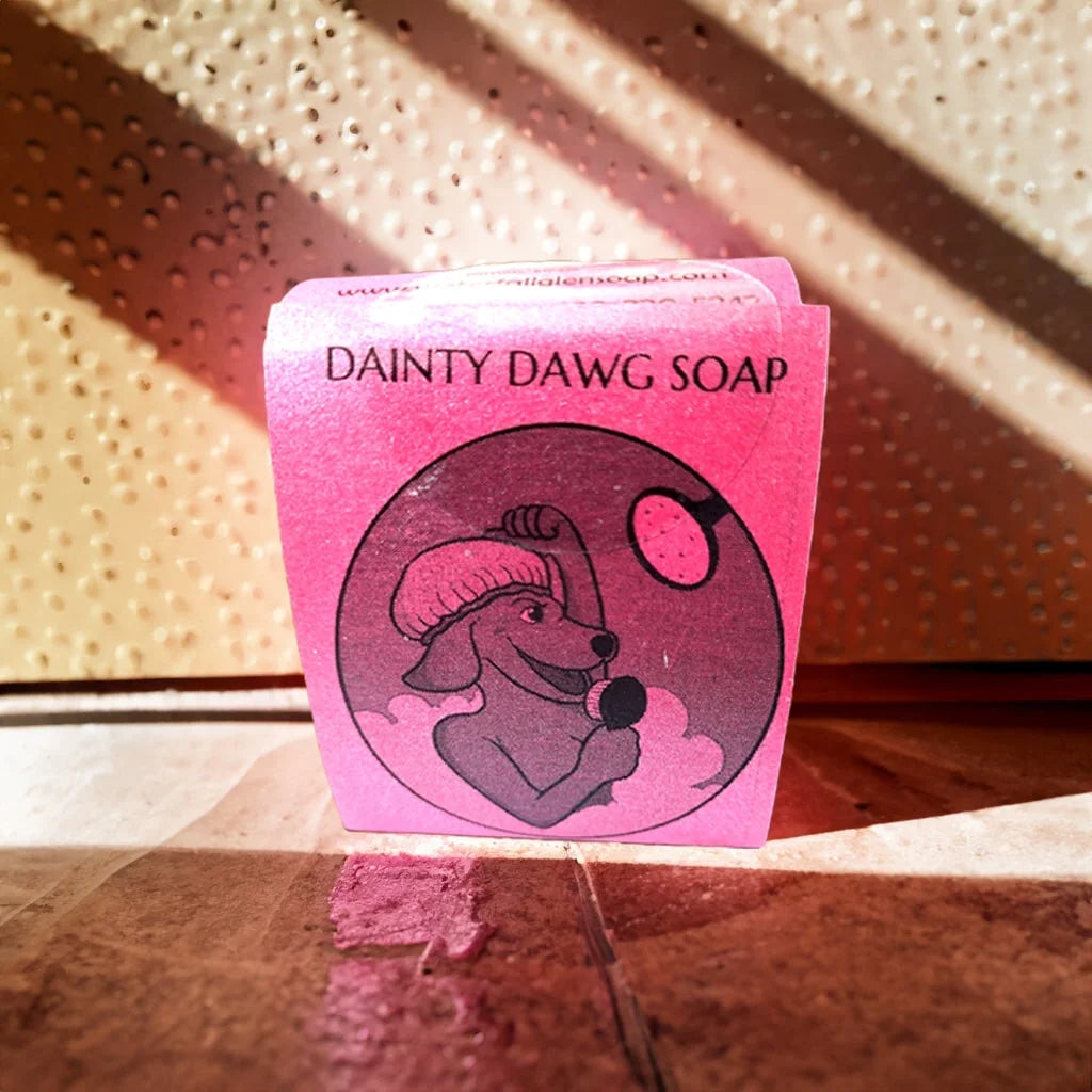 Dainty Dawg bar soap with a pink label of a cartoon dog washing itself and the bar soap is sitting on the bottom of a shower floor.
