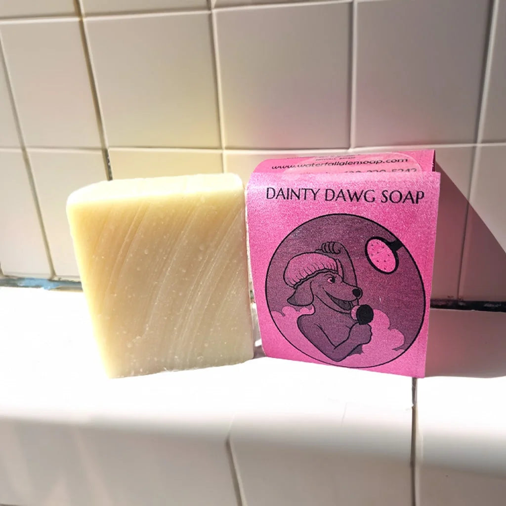 Dainty Dawg bar soap with a pink label of a cartoon dog washing itself and the bar soap is sitting on the bottom of a shower floor. With the bar soap with no label sitting next to it.