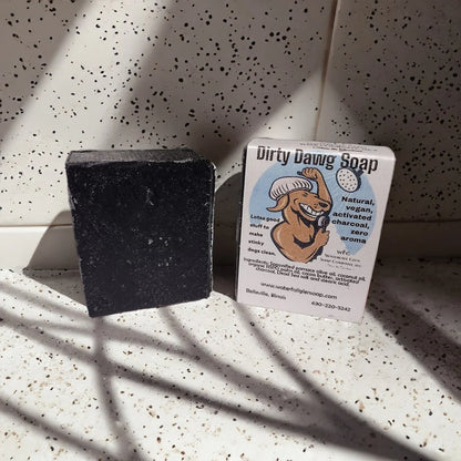 black bar of soap and its label dirty dawg soap next to it with a graphic of a dawg washing itself in the shower on the label. Both sitting on top of a shower floor.