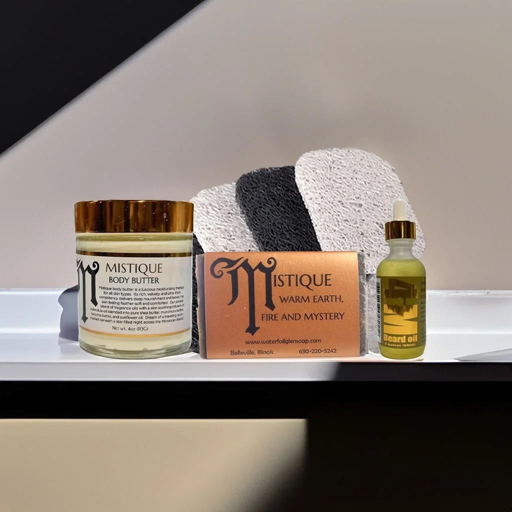 Mistique body butter on the left with a gold cap in a glass jar with a white label and gold reflective lettering and borders. In the middle is the Mistique bar soap with a orange label. On the right is the 4Him Beard Oil with a gold reflective label with the liquid inside a glass jar with a eyedropper tool. Behind these products is 4 soap saver bags. These are all sitting on top of a white table countertop.