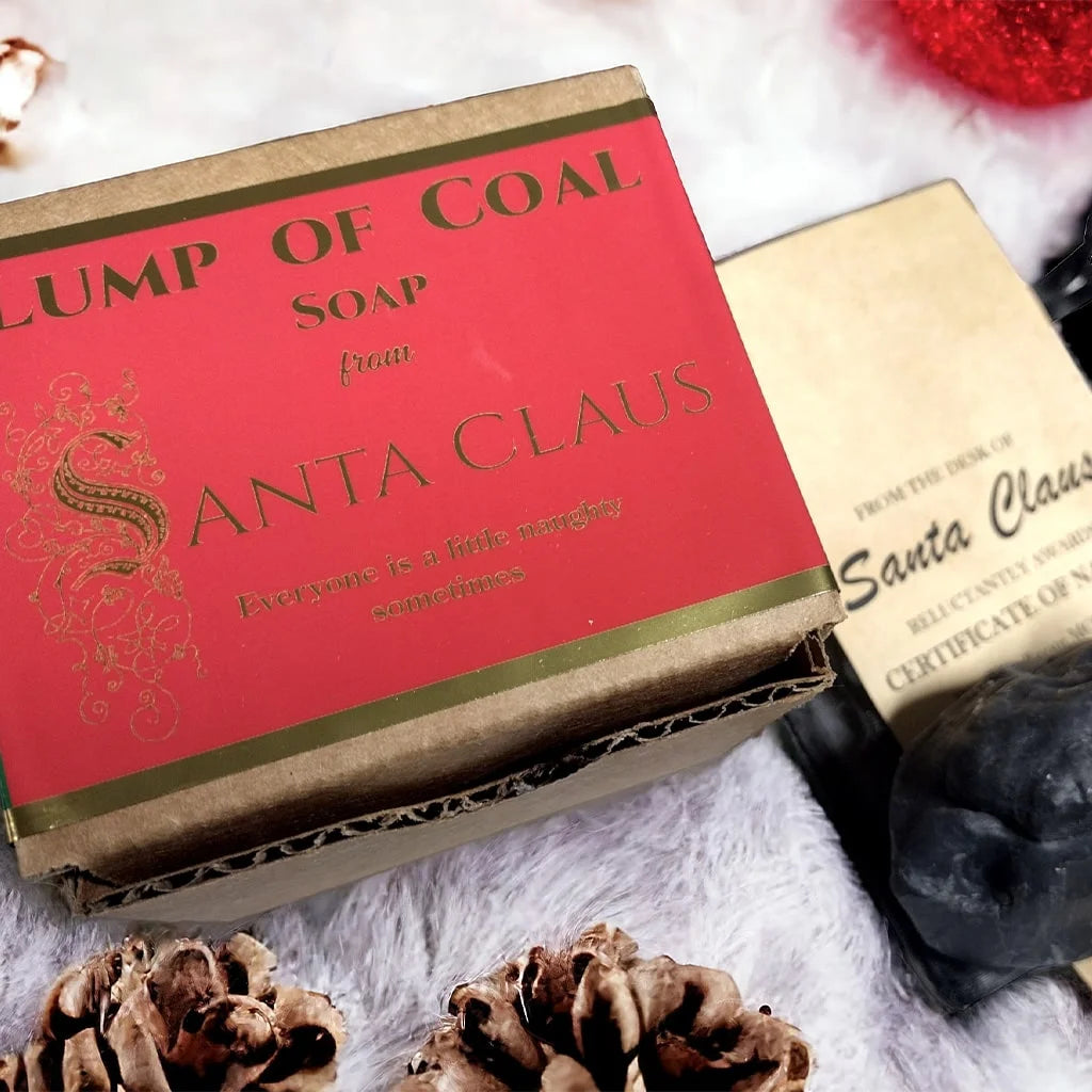 Lump of Coal square cardboard packing box with a red label and gold reflective lettering on top of a white carpet surrounded by ornaments, acorns and Santa's note.