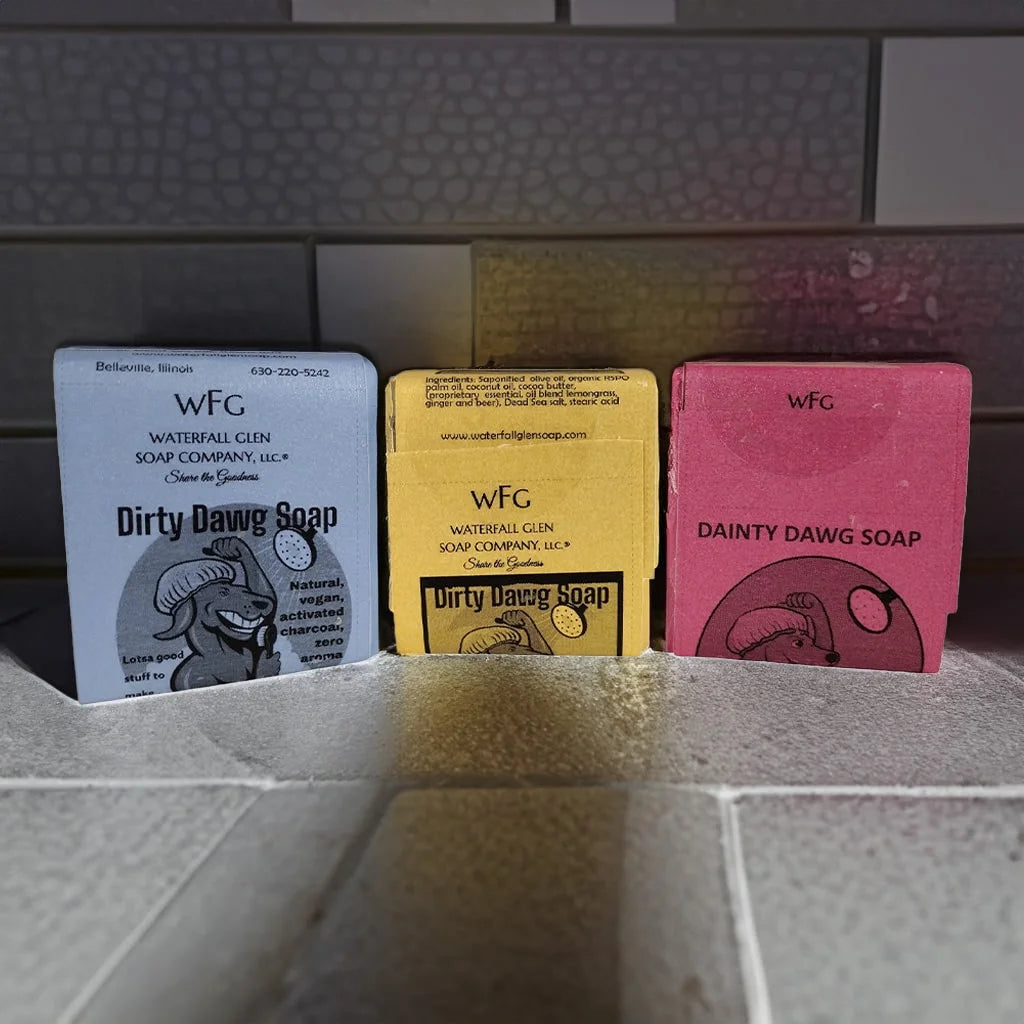 3 bar soaps sitting on the bottom of a shower floor. Each having a graphic of a cartoon dog washing itself. One soap has a blue label, the soap in the middle has a yellow label, the soap on the right has a red label.