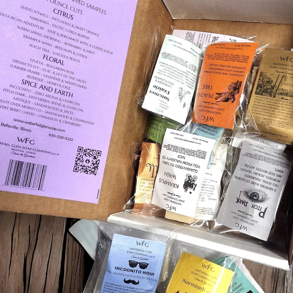Inside of a Sampler Box, the packaging is a cardboard box with a purple label and black text holding 15 different sample soaps ranging in colors from orange, yellow, black, purple and more.