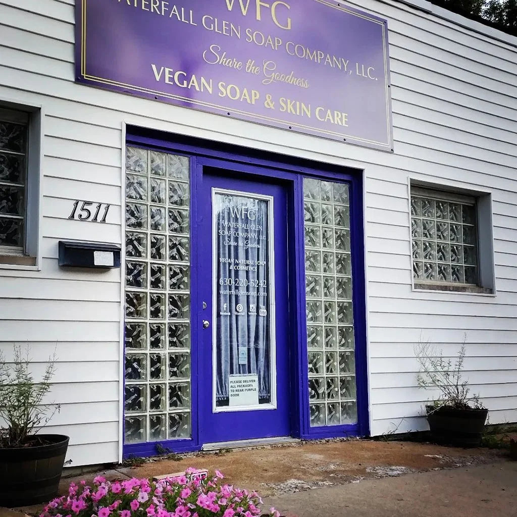 the front of the waterfall glen soap company building. A purple door with glass window side panels and a row of pink flowers in front of the concrete door step.