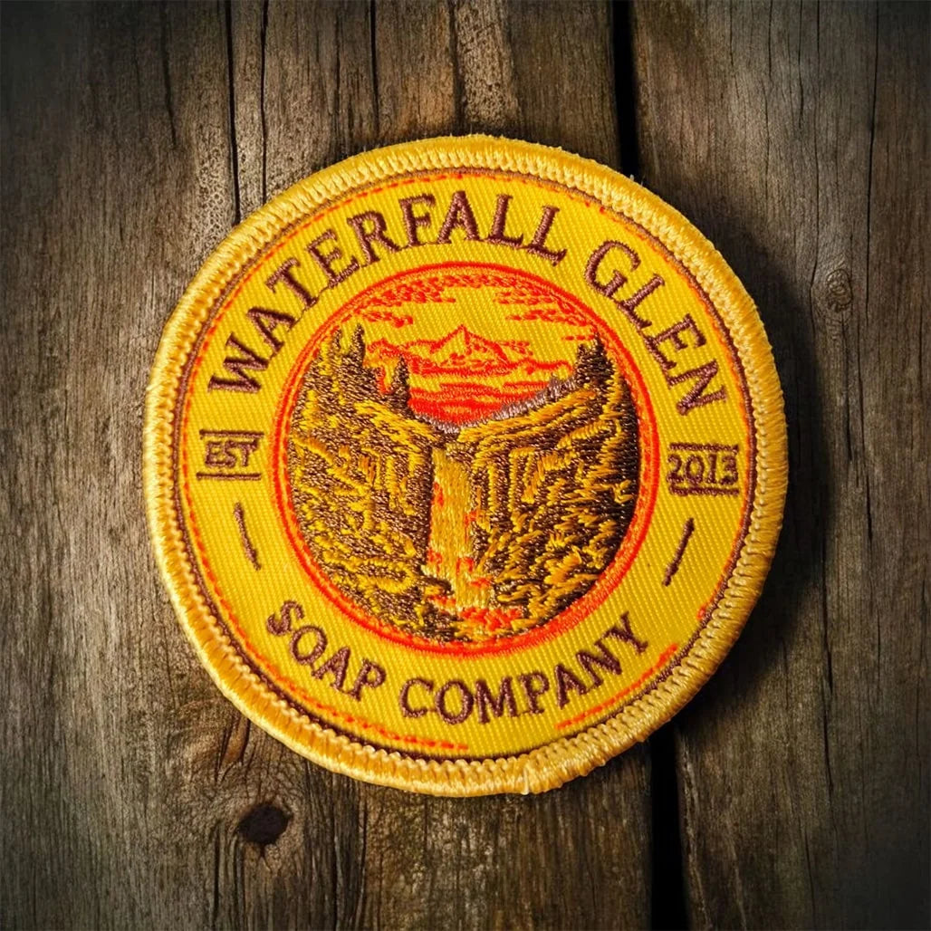 Yellow embroidered patch with company name and graphic in the middle of a waterfall and mountain and sunset.
